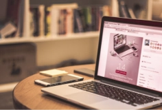 A laptop with a smartphone on the desk and books on a shelf.